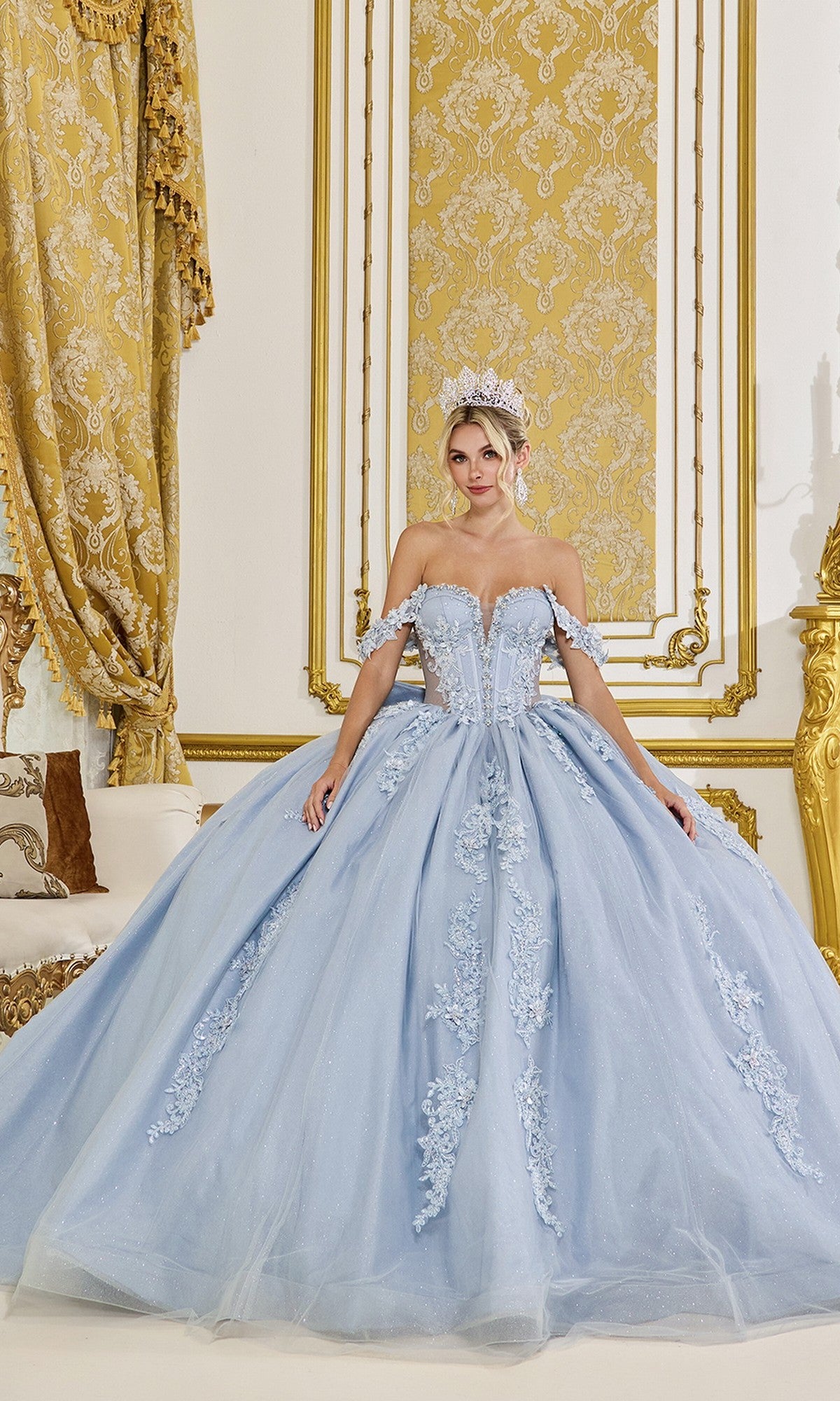 A woman wearing the Quinceanera Dress 5088 By Dancing Queen in dusty blue, featuring an off-the-shoulder lace corset bodice and voluminous skirt, poses with both hands on her dress in an ornate gold-accented room.